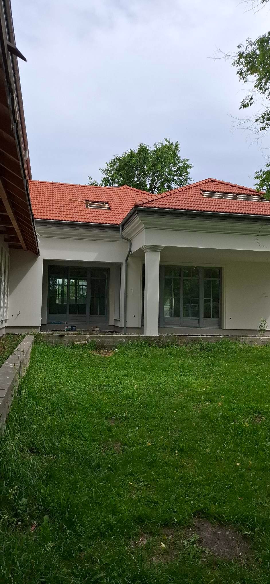 Dom z jasną elewacją i czerwoną dachówką, widok z zewnątrz na trawnik. Szare, duże okna i kolumny przy wejściu. Częściowo widoczne orynnowanie.