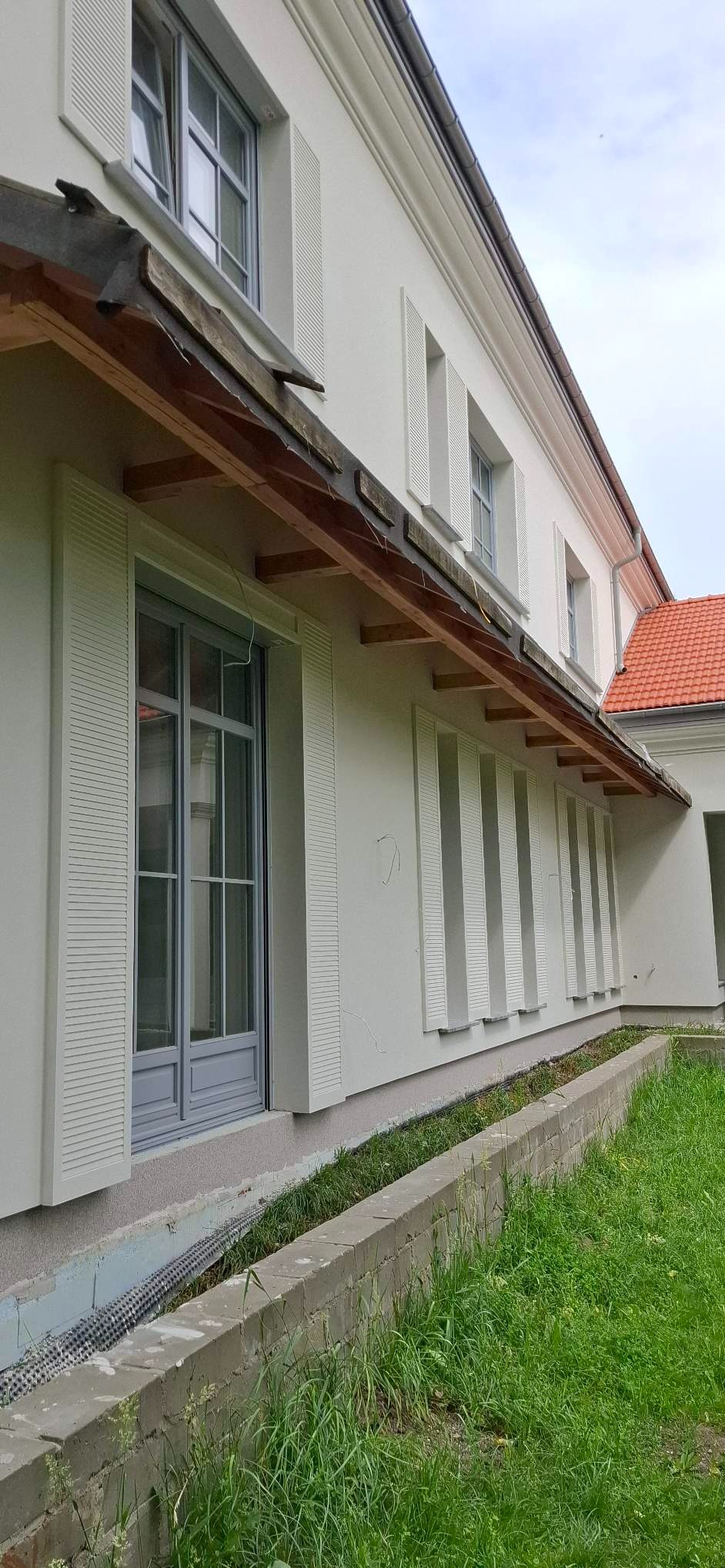 Jasna elewacja budynku z szarymi detalami okiennymi i dekoracyjnymi panelami. Widoczne elementy drewniane pod dachem i fragment dachu z czerwonej dachówki.