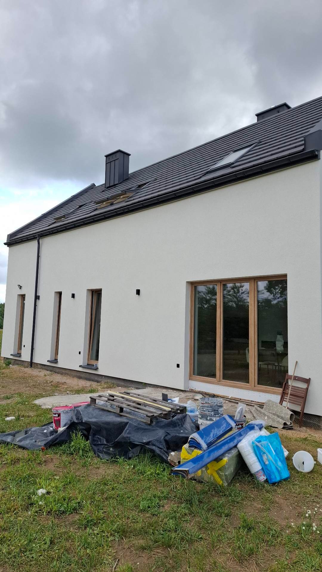 Otynkowana ściana domu jednorodzinnego w Wołominie. Widoczne okna z drewnianą ramą, ciemna dachówka i przygotowane materiały budowlane.