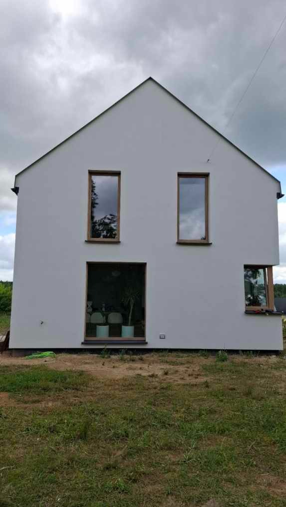 Nowoczesny dom z białą elewacją i drewnianymi oknami, otoczony zielenią. Duże okno tarasowe z widokiem na wnętrze. Minimalistyczny design.