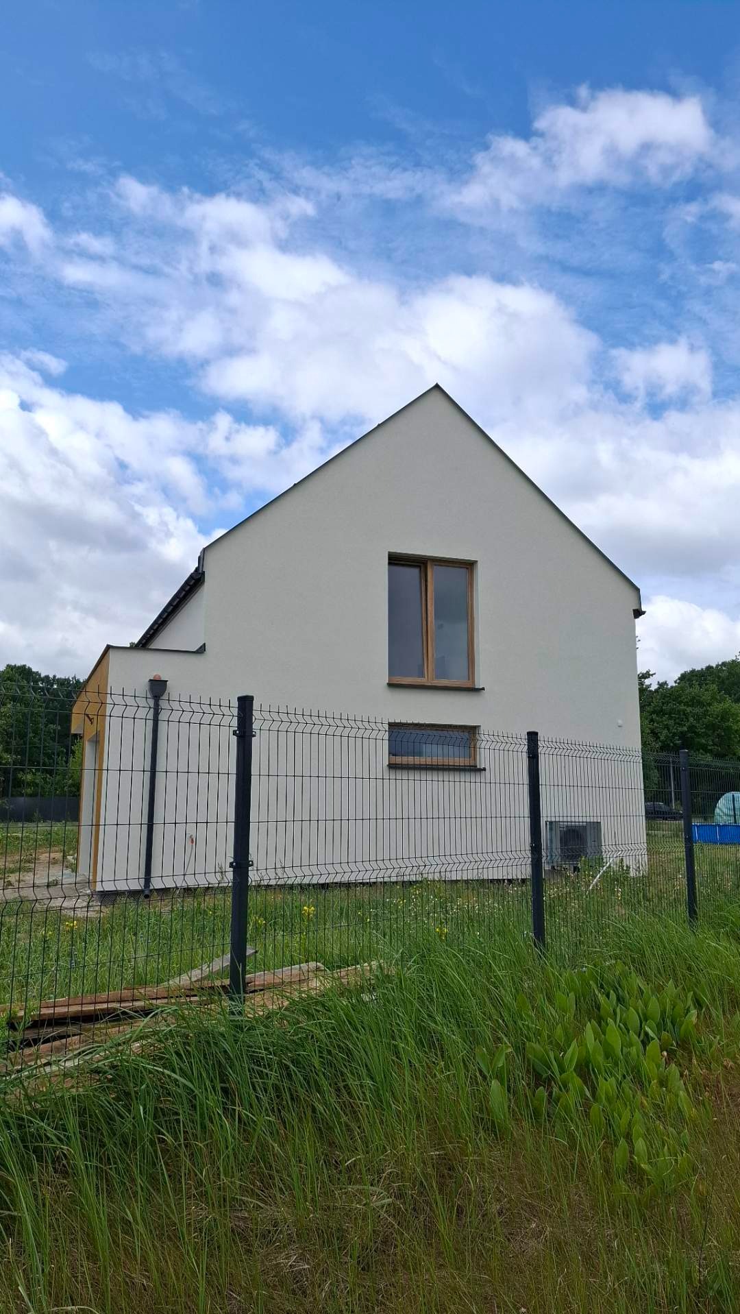 Nowoczesny, otynkowany dom jednorodzinny z drewnianą stolarką okienną, ogrodzony metalową siatką na tle błękitnego nieba z chmurami.