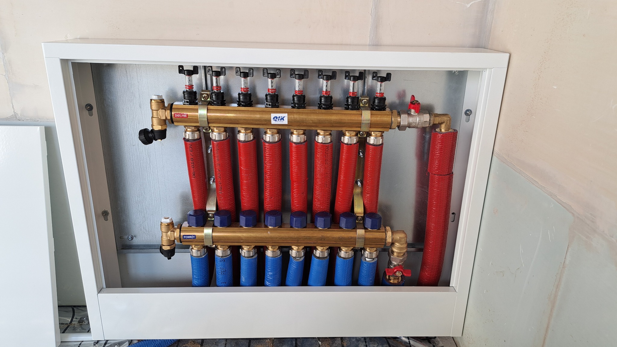 Rozdzielacz ogrzewania podłogowego w białej szafce, z czerwonymi i niebieskimi rurami, zamontowany na ścianie w trakcie prac wykończeniowych.