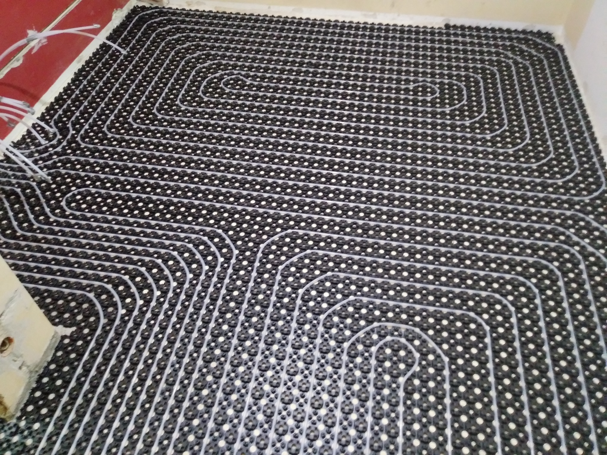 Czarna mata z wypustkami i ułożone na niej białe rurki ogrzewania podłogowego, tworzące równoległe, regularne linie. Widoczne fragmenty ściany i podłączenia rur.