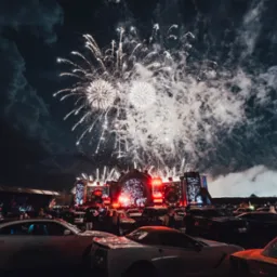 Nocny widok na scenę podczas wydarzenia plenerowego z efektownymi fajerwerkami nad samochodami. W tle ciemne chmury i oświetlona scena.