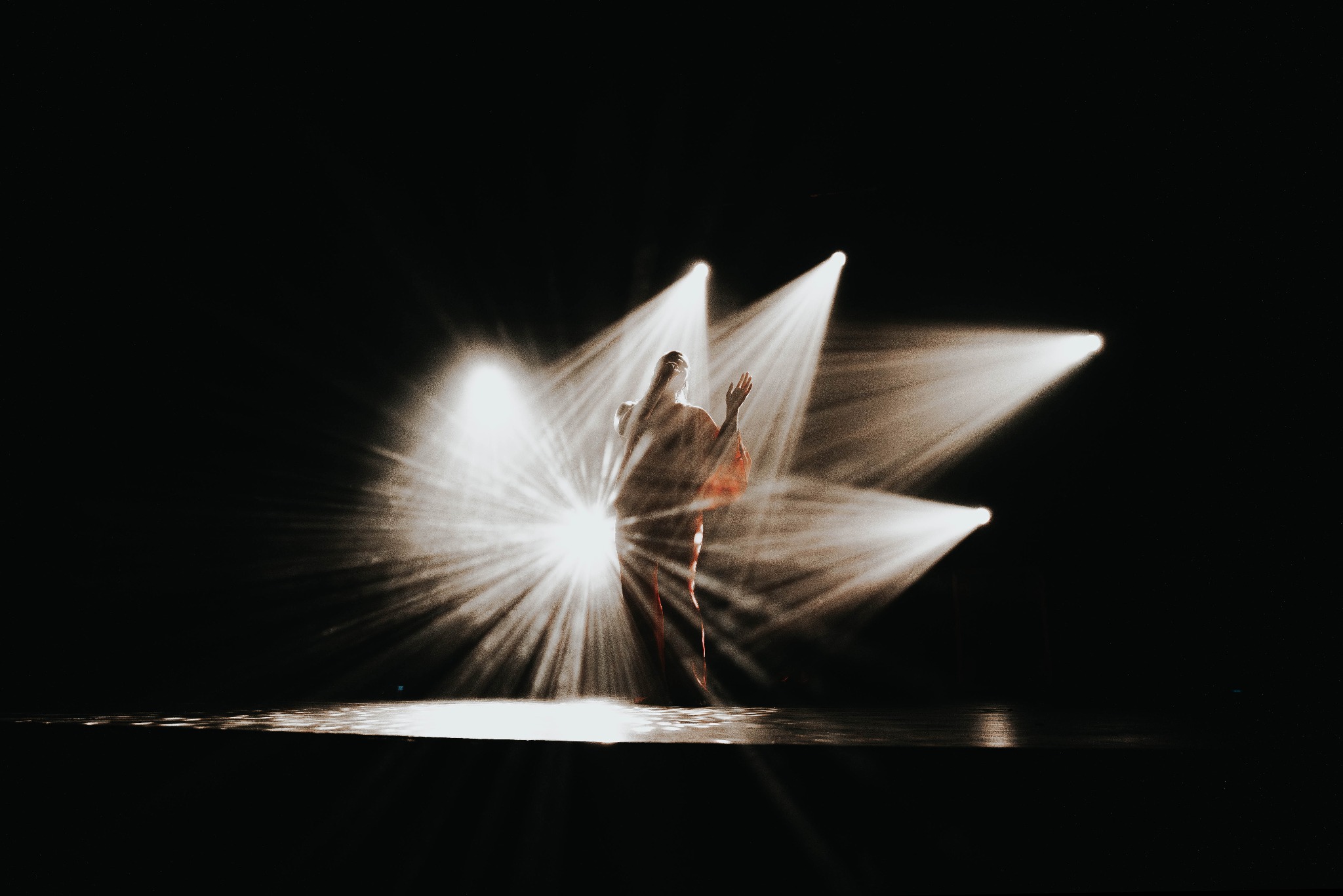 Sylwetka tancerki oświetlona snopami światła na scenie. Ujęcie w kontraście, z dominującą czernią tła i jasnymi promieniami reflektorów.