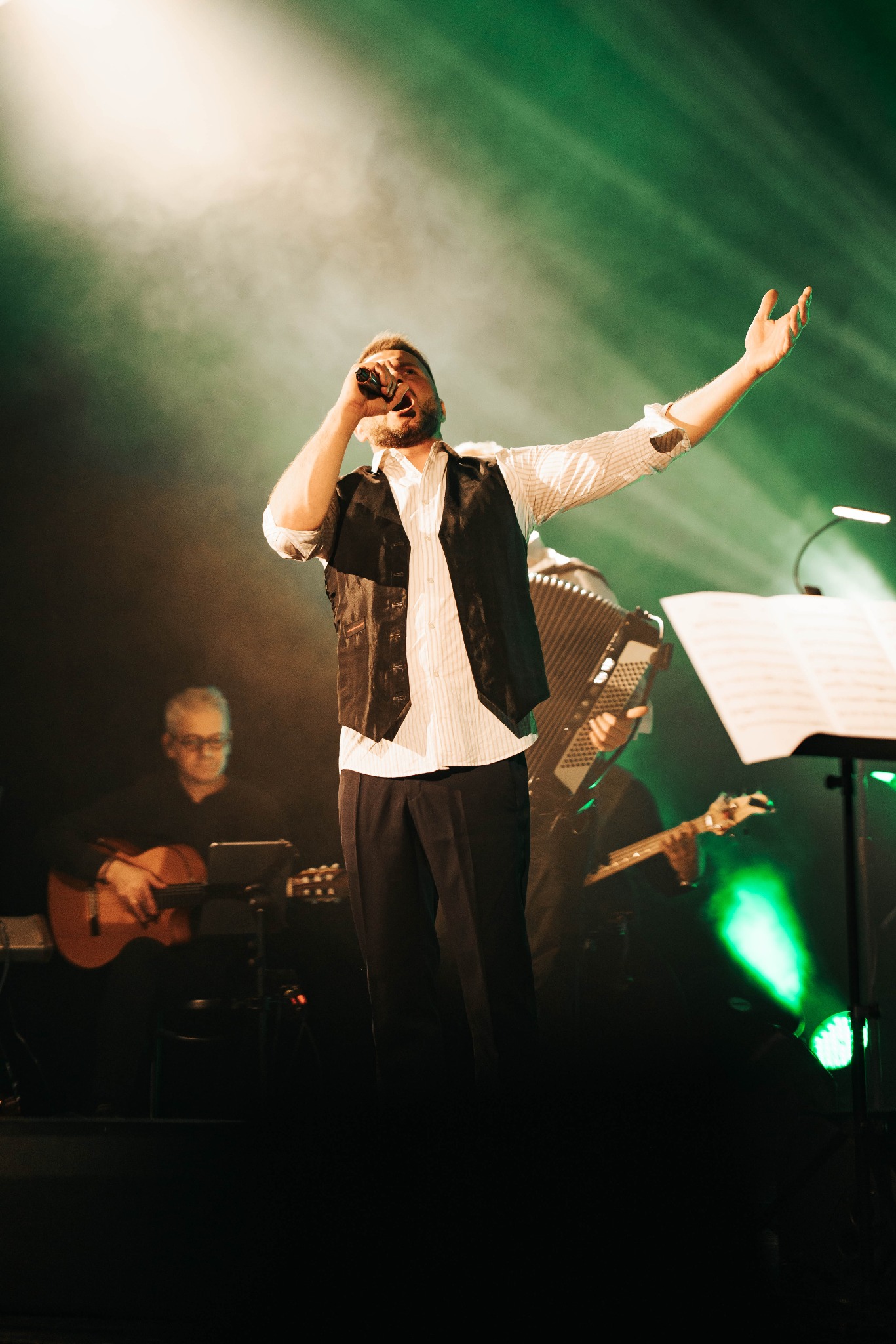 Wokalista w kamizelce śpiewa do mikrofonu na scenie w zielonym oświetleniu, w tle muzycy z gitarą i akordeonem. Artysta z uniesioną ręką.