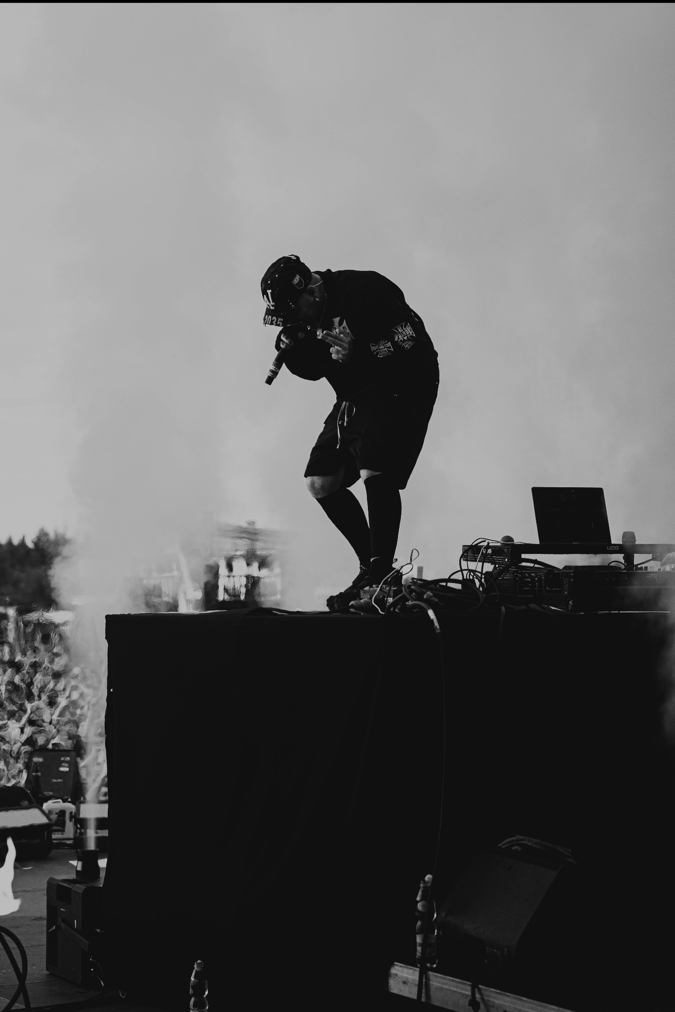 Czarno-białe ujęcie rapera na scenie podczas występu, z mikrofonem w dłoni i laptopem obok, publiczność w tle, dynamiczne oświetlenie.