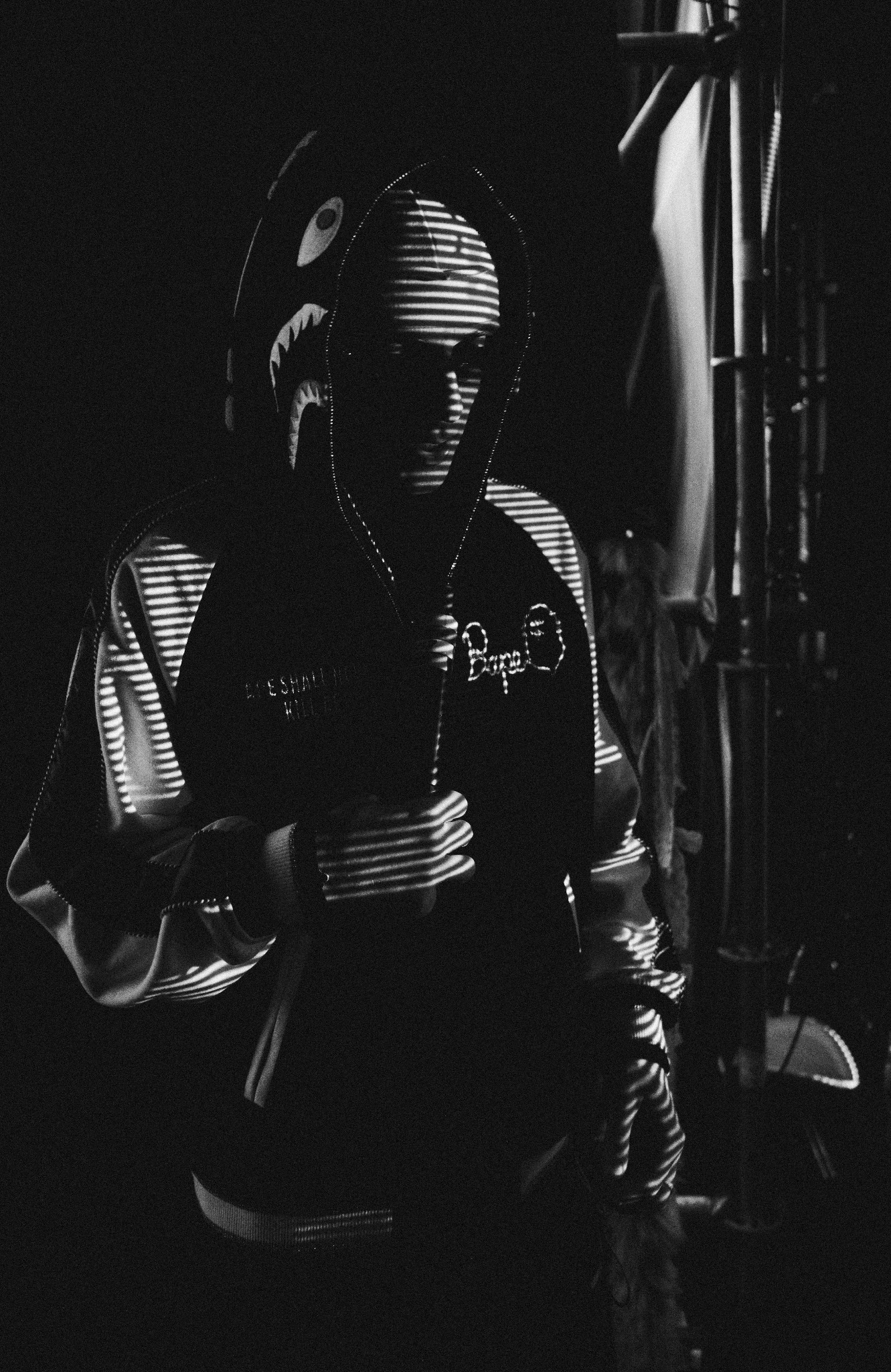 Czarno-białe zdjęcie osoby w bluzie z kapturem w stylu BAPE, oświetlonej przez światło padające przez żaluzje, tworzące unikalny efekt cieniowania na twarzy i ubraniu, w tle widoczne elementy...