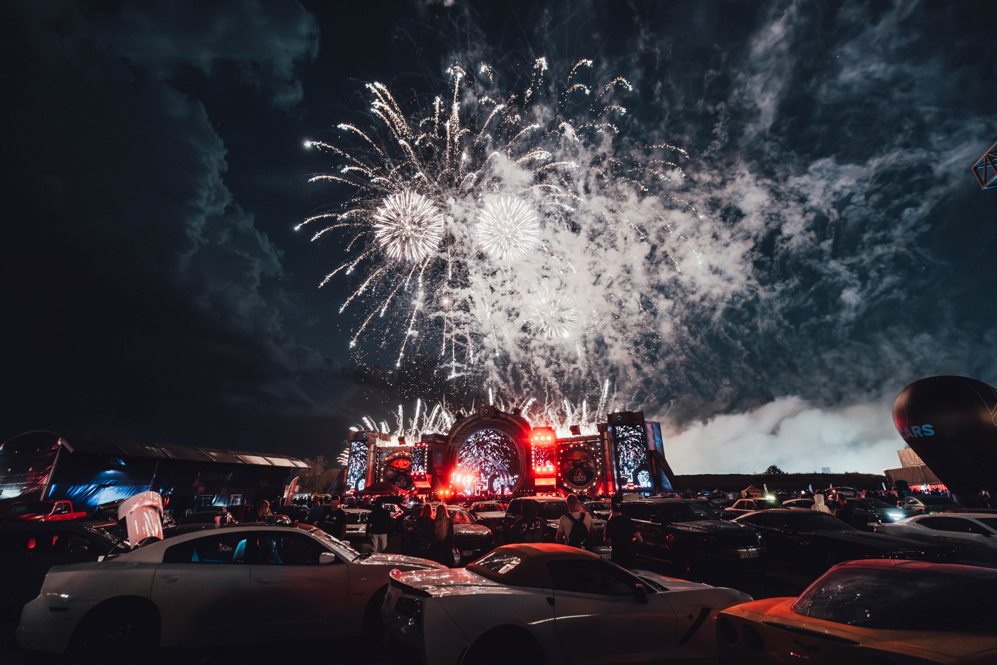 Nocny widok na scenę podczas wydarzenia plenerowego z efektownymi fajerwerkami nad samochodami. W tle ciemne chmury i oświetlona scena.
