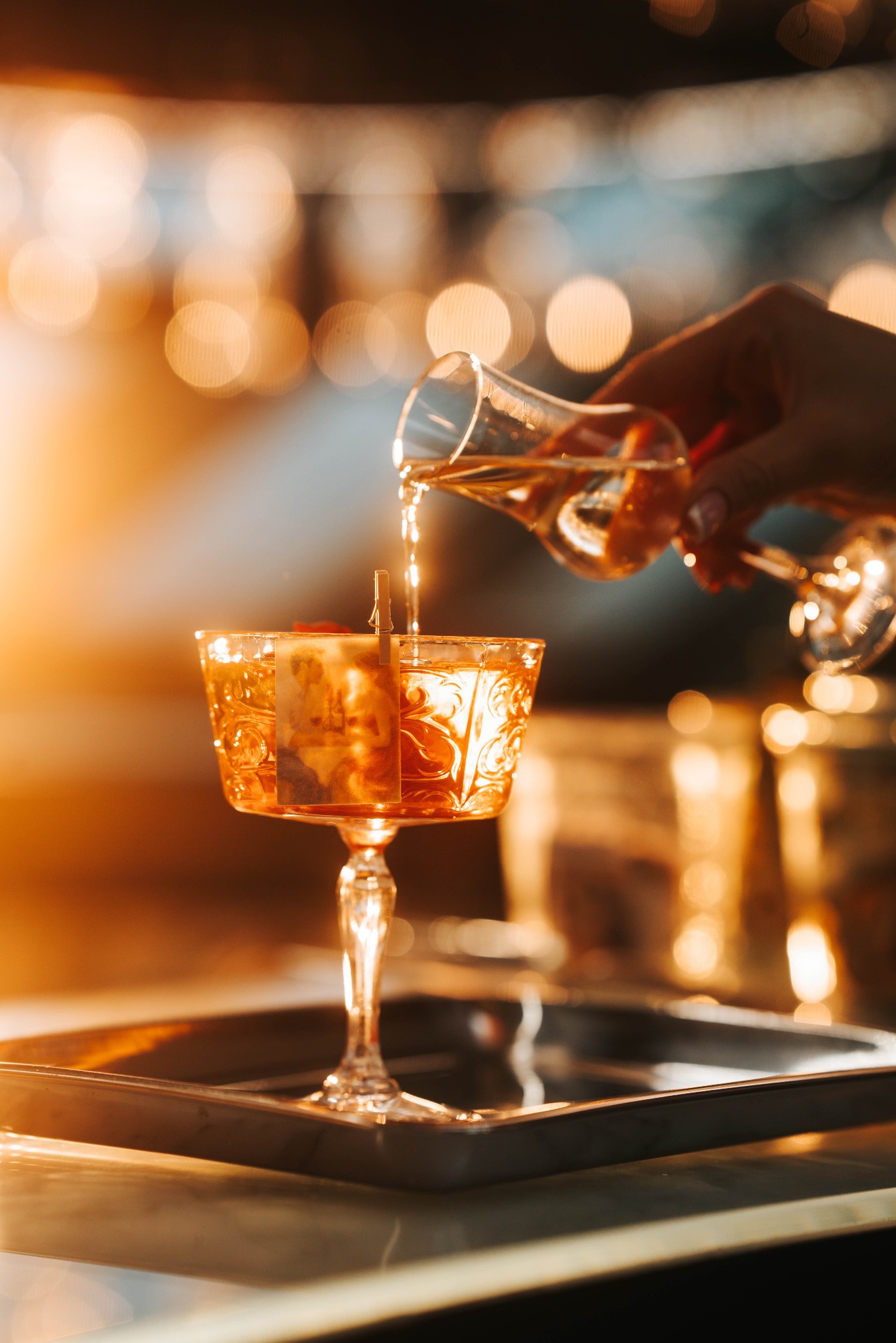 Barman nalewa drink do ozdobnego kieliszka z przymocowanym zdjęciem, ciepłe światło i bokeh w tle, na srebrnej tacy.