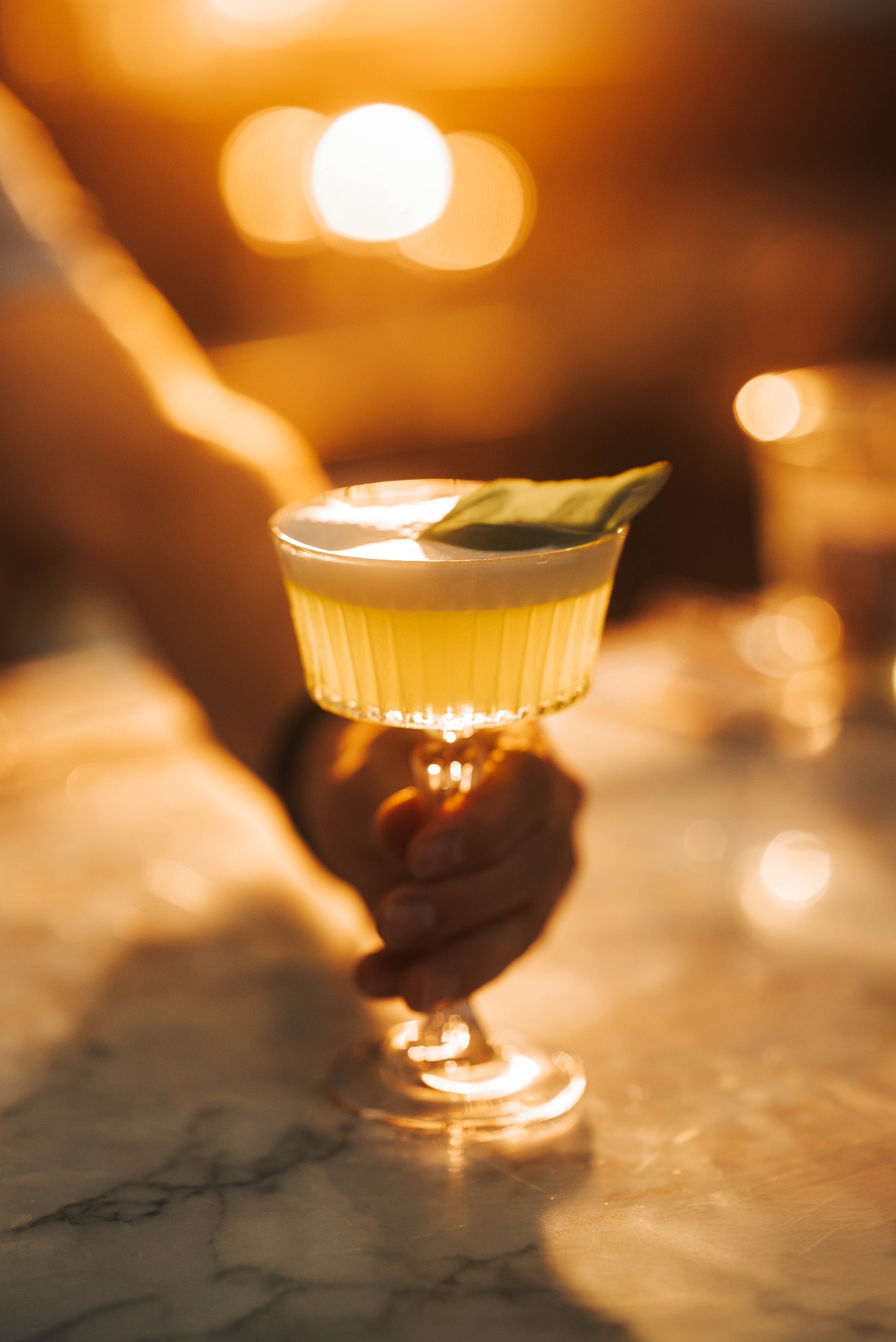 Dłoń trzyma kieliszek z żółtym koktajlem, udekorowanym listkiem. Ciepłe światło, marmurowy blat, atmosfera baru.