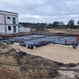 DFDN SP. Z O.O. - Fundamenty domu w trakcie budowy: zbrojenie, izolacja, pionowe rury. W tle ściany parteru z pustaków i materiały budowlane. Pochmurne niebo.