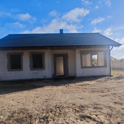 DFDN SP. Z O.O. - Niedokończony dom z białych bloczków, z czarnym dachem, bez okien i drzwi, na tle błękitnego nieba z chmurami i jesiennego krajobrazu.