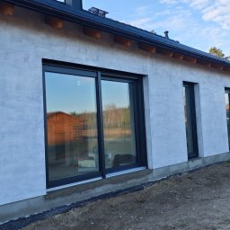 DFDN SP. Z O.O. - Boczny widok domu z szarą, teksturowaną elewacją i czarnymi ramami okiennymi. Widoczny fragment dachu, rynna oraz osoba w oddali. W oknach odbija się krajobraz.