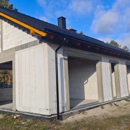 DFDN SP. Z O.O. - Niedokończony dom z białych bloczków, czarny dach z żółtymi elementami, widoczne otwory okienne i drzwiowe, teren budowy, błękitne niebo z chmurami w tle.