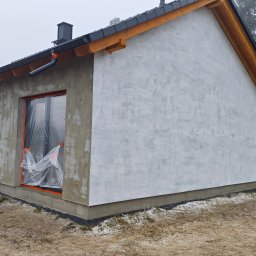 DFDN SP. Z O.O. - Niedokończony dom z oknami zabezpieczonymi folią, ściany częściowo pokryte białą warstwą, widoczne drewniane elementy dachu i szara elewacja. W tle las i samochód.