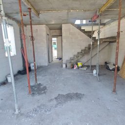 DFDN SP. Z O.O. - Wnętrze domu w stanie surowym: ściany z bloczków, betonowe schody, podpory stropowe. Widoczne okno i przejścia między pomieszczeniami. Podłoże betonowe.