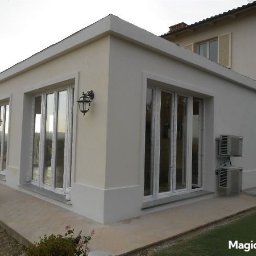 Ecohome24 sp. z o.o. - Jasny, parterowy budynek z dużymi oknami i zewnętrznymi lampami, widoczne klimatyzatory na ścianie. Budynek otoczony zielenią, w tle wzgórza.