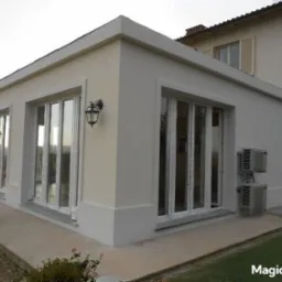 Jasny, parterowy budynek z dużymi oknami i zewnętrznymi lampami, widoczne klimatyzatory na ścianie. Budynek otoczony zielenią, w tle wzgórza.