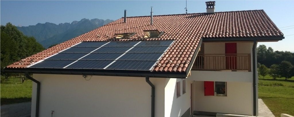 Jasny dom z panelami słonecznymi na dachu z dachówką, na tle gór i zieleni. Widok z boku, w słoneczny dzień. Czerwone akcenty: drzwi i okiennice.