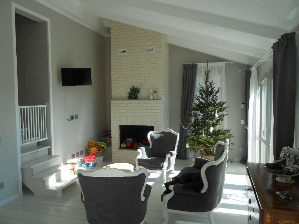 Jasne wnętrze domu z kominkiem z cegły, choinką i eleganckimi fotelami. Widoczne schody z barierką i telewizor na ścianie. Wnętrze w neutralnej kolorystyce.