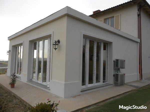 Jasny, parterowy budynek z dużymi oknami i zewnętrznymi lampami, widoczne klimatyzatory na ścianie. Budynek otoczony zielenią, w tle wzgórza.