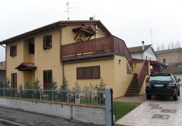 Żółty dom z drewnianym balkonem i schodami zewnętrznymi, widok z ulicy. Metalowe ogrodzenie i małe drzewka przed budynkiem. Czarny samochód zaparkowany obok schodów.