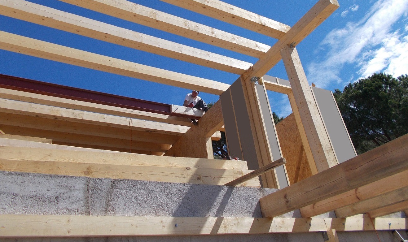 Wnętrze w trakcie budowy: drewniana konstrukcja szkieletowa domu z widocznym pracownikiem na tle błękitnego nieba i drzew. Fragment ściany z płyt OSB i szarymi panelami.