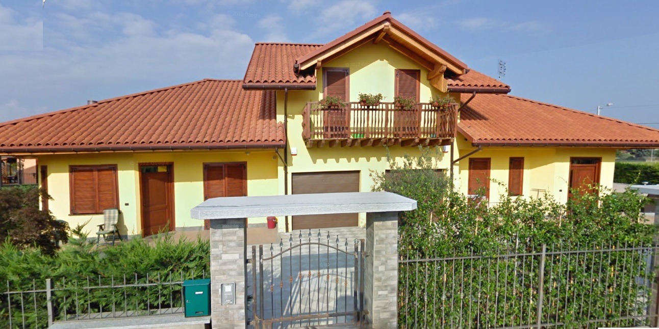 Żółty dom z brązowym dachem i balkonem z kwiatami, widoczny zza ogrodzenia. Dom jednorodzinny z garażem i drewnianymi okiennicami w słoneczny dzień.