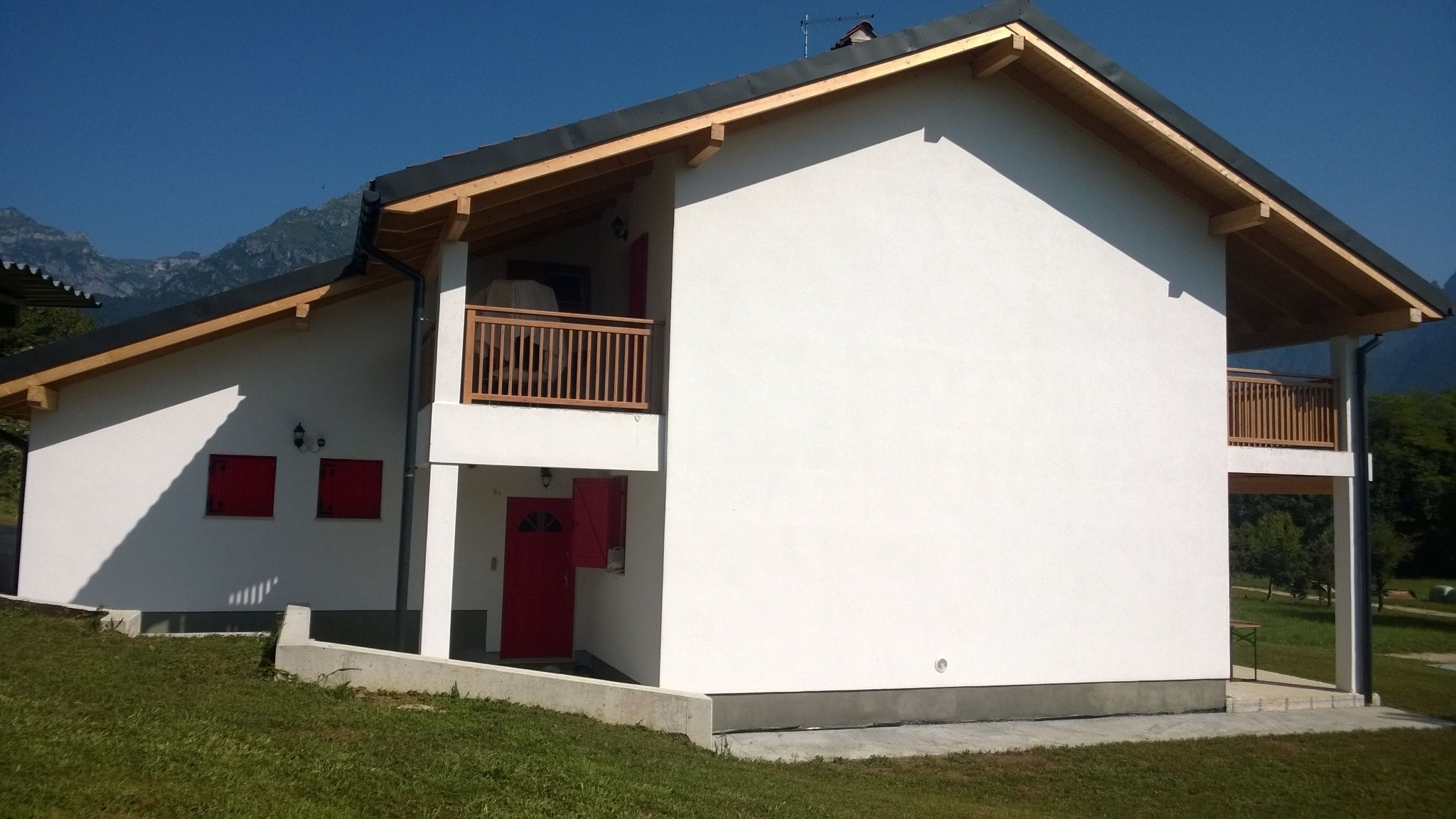 Dwukondygnacyjny dom z białymi ścianami, drewnianą konstrukcją dachu i czerwonymi akcentami okien i drzwi, na tle górskiego krajobrazu w słoneczny dzień.