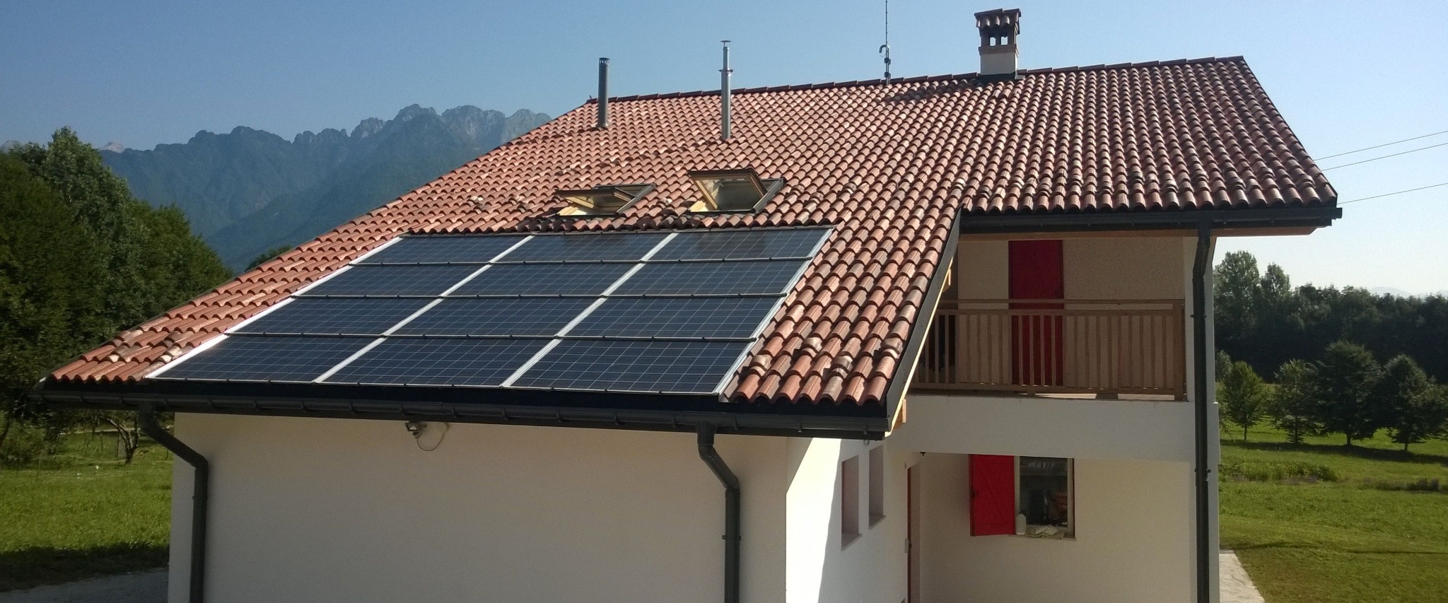 Gotowy dom z panelami słonecznymi na dachu z dachówką ceramiczną, w tle góry i las. Widok na balkon z czerwonymi drzwiami.