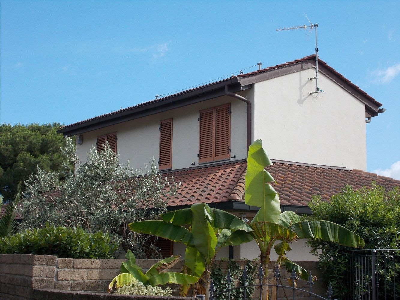 Dwukondygnacyjny dom z jasną elewacją i brązową dachówką, widoczny zza zieleni. Antena na dachu. Okiennice w kolorze brązu.