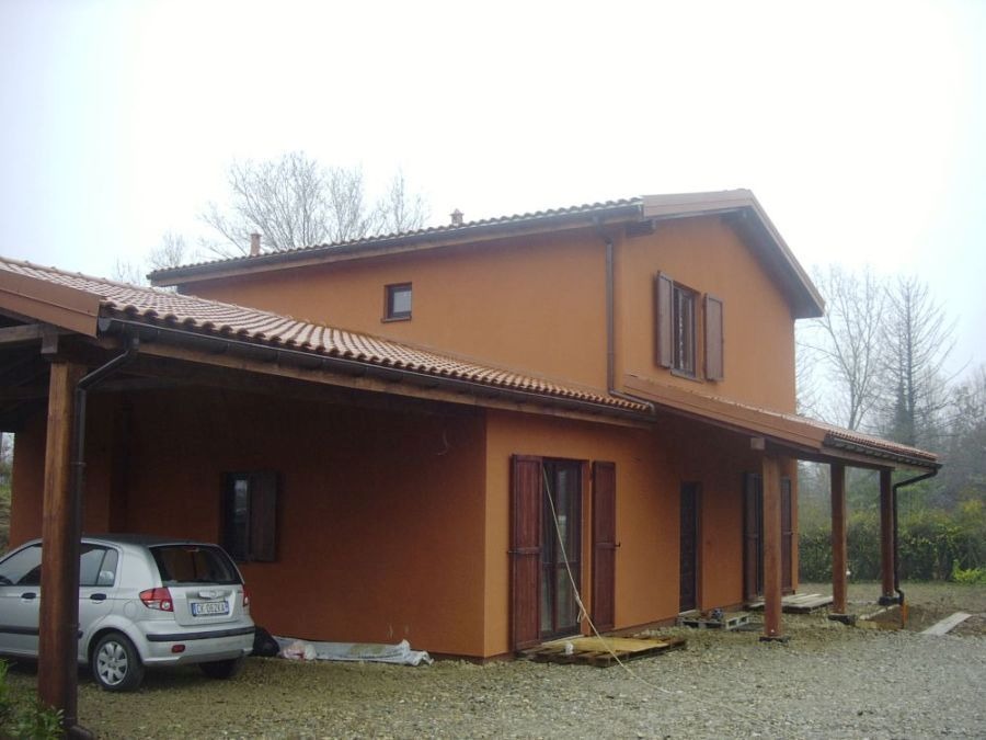 Gotowy dom z pomarańczową elewacją i zadaszonym miejscem parkingowym. Widoczne okiennice i dachówka. Na podjeździe stoi srebrny samochód.