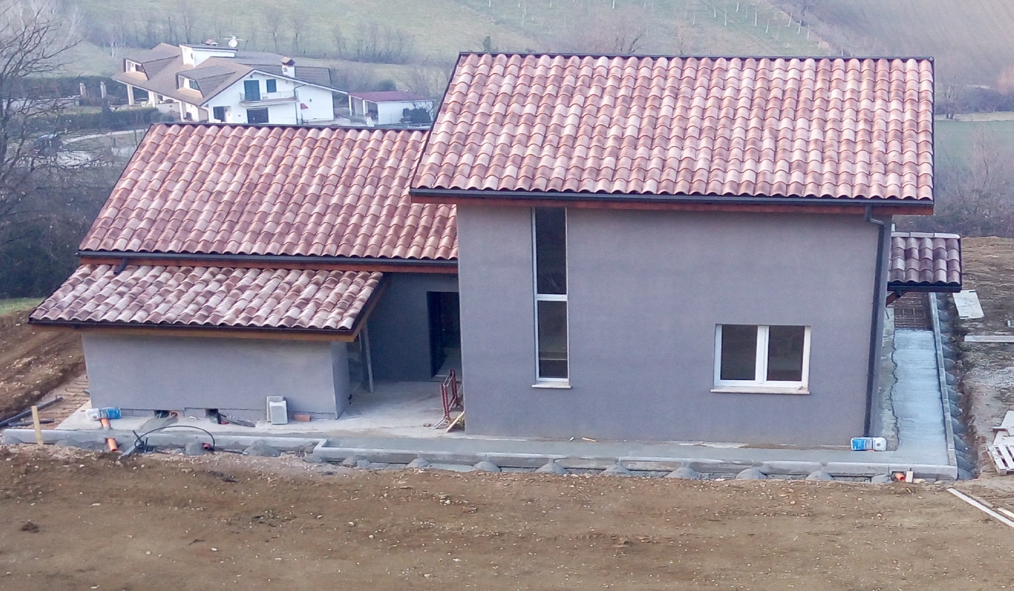 Szary dom z czerwoną dachówką w trakcie budowy, widok z zewnątrz. Widoczne fundamenty i otoczenie placu budowy. W tle zabudowa mieszkalna i wzgórza.