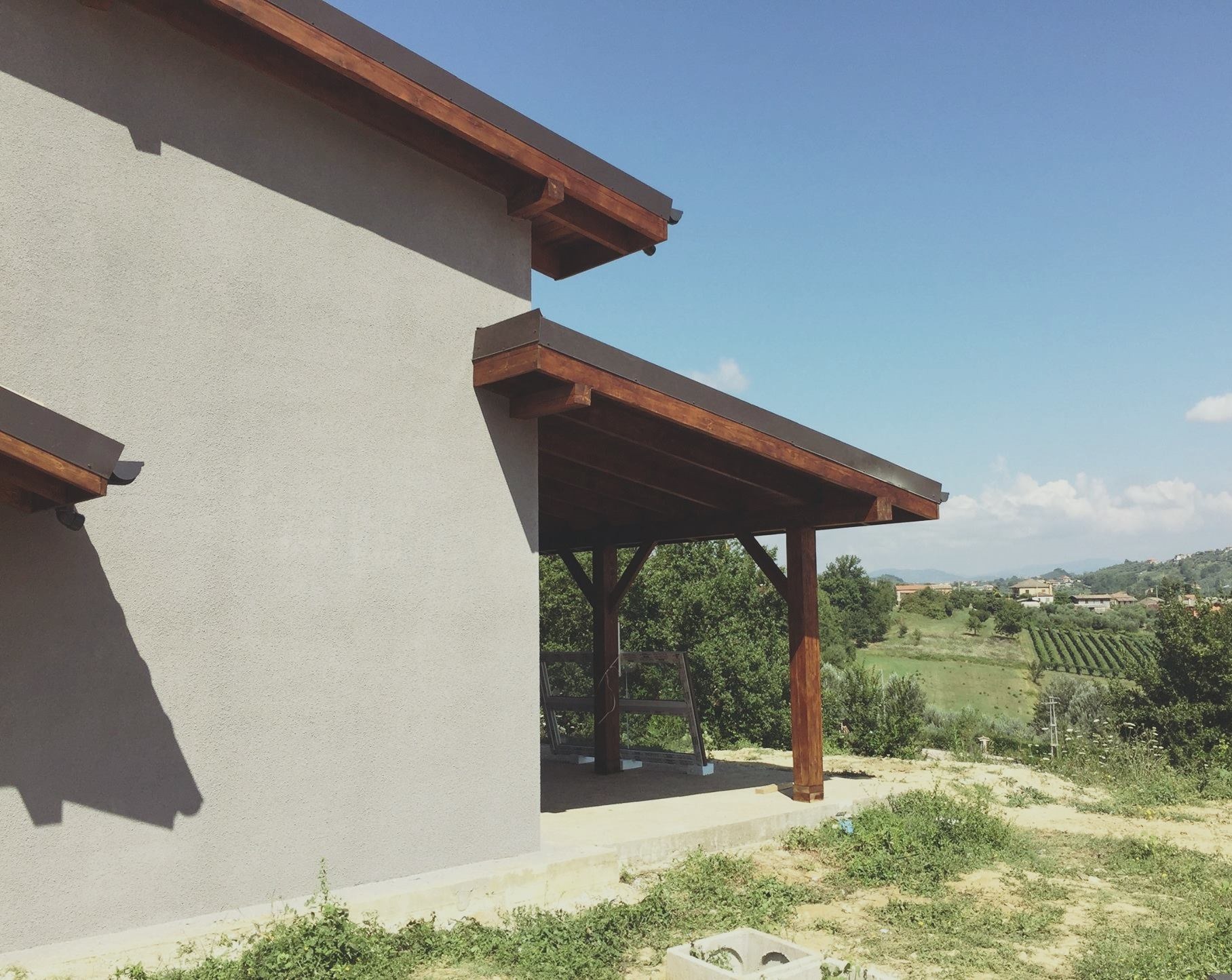 Nowoczesny dom z szarą elewacją i drewnianą konstrukcją zadaszenia, na tle zielonego krajobrazu z widocznymi wzgórzami i błękitnym niebem.