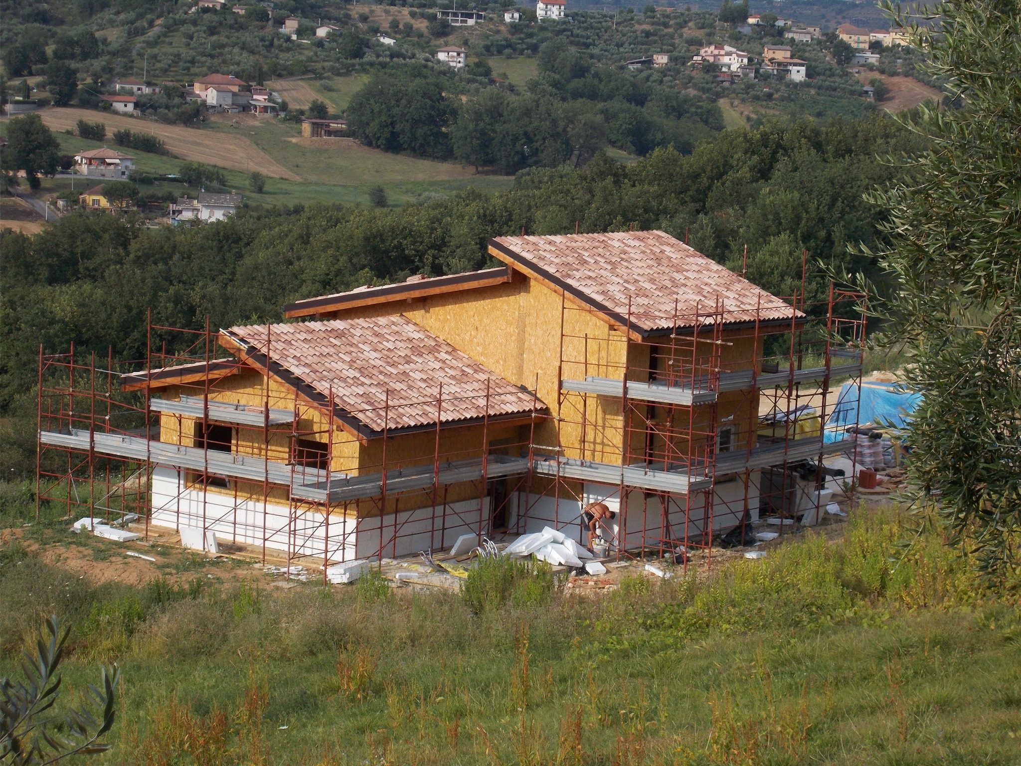 Wznoszenie dwukondygnacyjnego domu szkieletowego na tle malowniczego krajobrazu z rusztowaniami i częściowo ułożoną dachówką. Widoczne białe płyty izolacyjne.