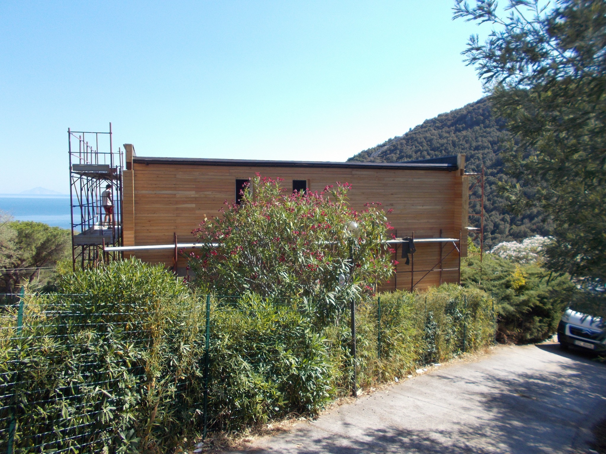Wznoszony dom z drewnianą elewacją, otoczony zielenią. Na rusztowaniu stoi osoba. W tle widok na morze i wzgórza. Słoneczny dzień.