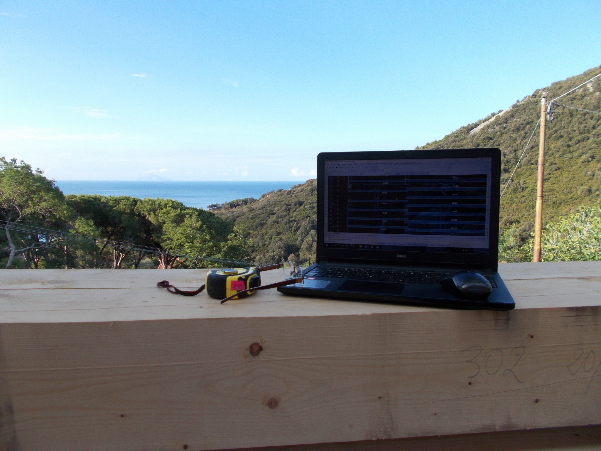 Laptop z planami na drewnianej belce, miernik i okulary, w tle malowniczy krajobraz z morzem i wzgórzami. Koncepcja projektowania domów z naturalnych materiałów.