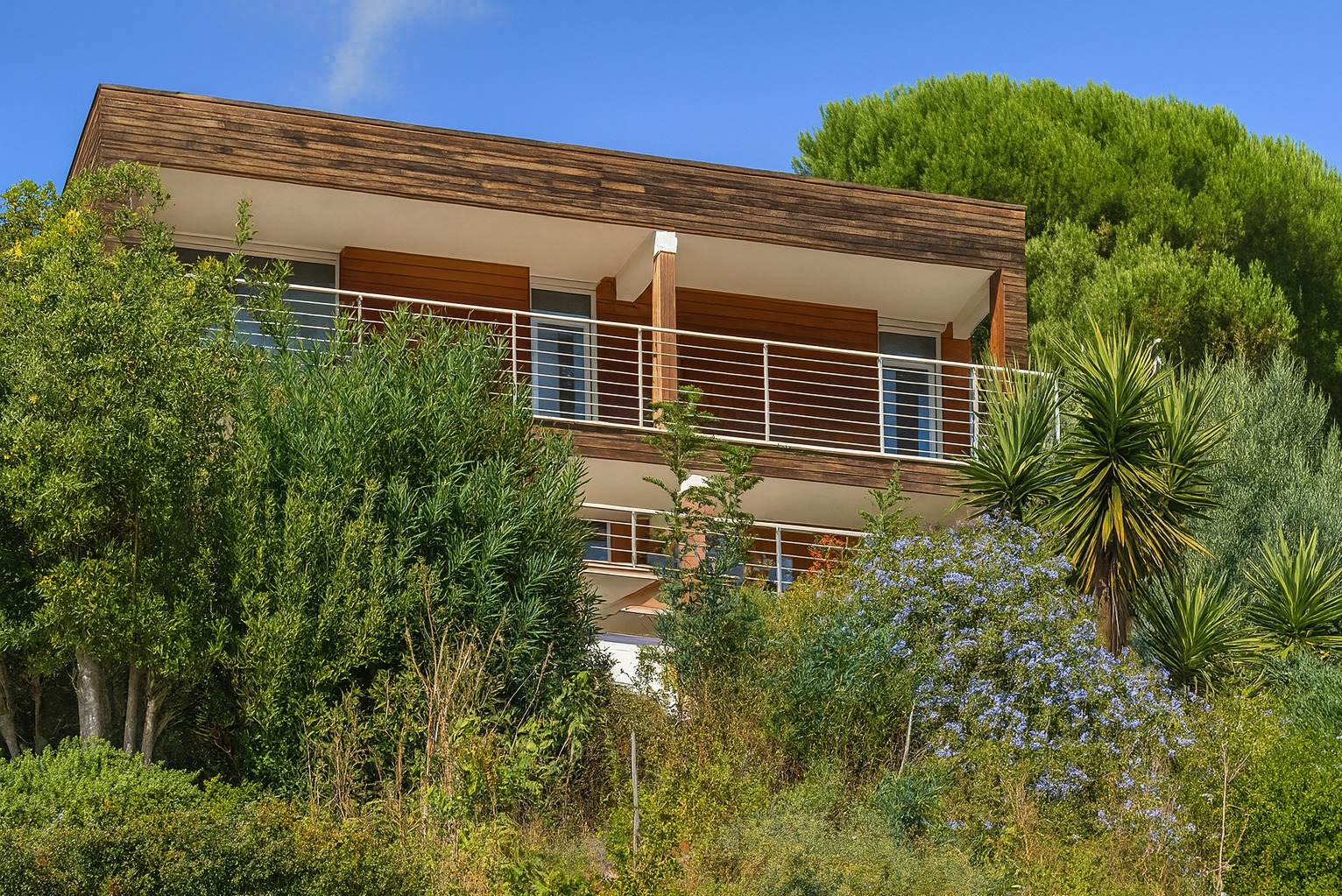 Dwukondygnacyjny dom z drewnianą elewacją i balkonami, otoczony bujną roślinnością w słoneczny dzień, prezentujący nowoczesny styl architektoniczny.