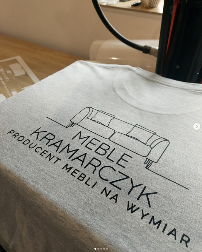 Szara koszulka z czarnym nadrukiem logotypu 'MEBLE KRAMARCZYK', grafika sofy. Nadruk wykonany metodą folii flex, widoczna prasa termotransferowa w tle.