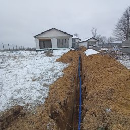 Piotr Gańko - Wykop z niebieską rurą kablową na tle budowy domu w zimowej scenerii. Widok z góry na wykopaną ziemię, rurę i budynek w stanie surowym otwartym.