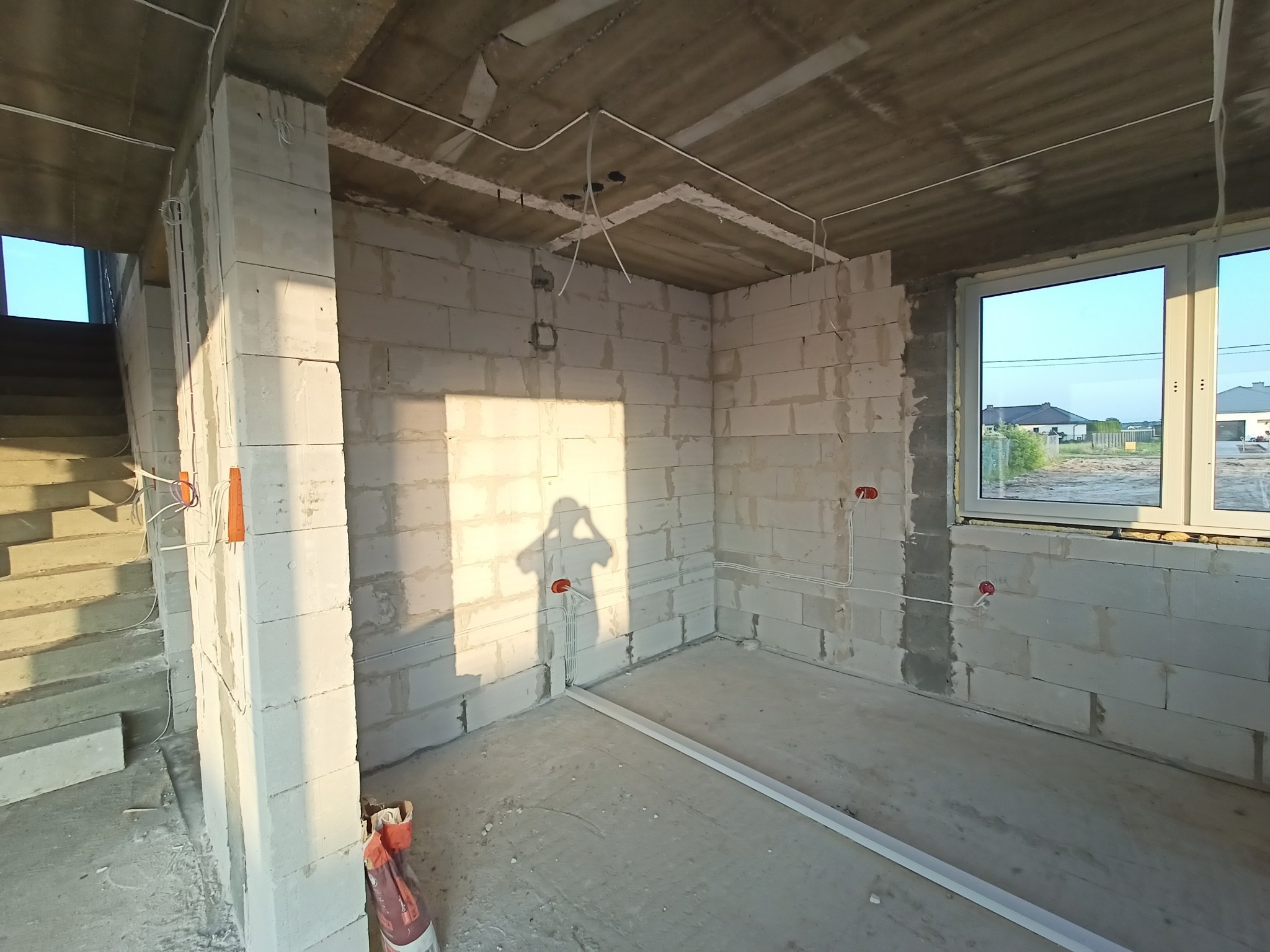 Wnętrze w stanie surowym z widoczną instalacją elektryczną: kable poprowadzone po ścianach z betonu komórkowego i suficie. Widoczne puszki elektryczne i cień osoby.