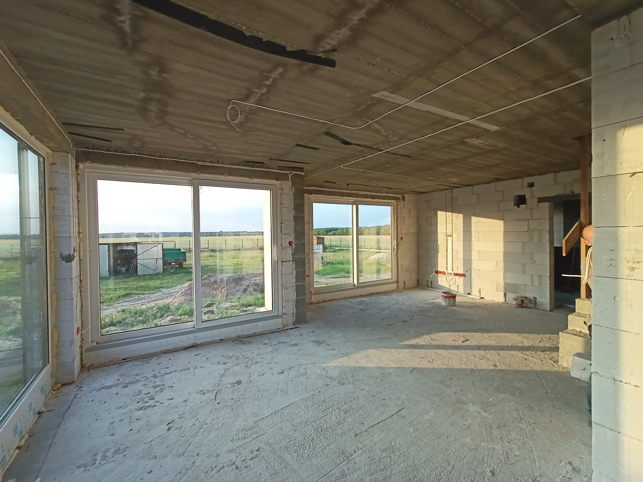 Surowy stan wnętrza domu w trakcie budowy: widoczne okna, ściany z betonu komórkowego i rozprowadzone kable elektryczne pod sufitem. W tle zieleń.