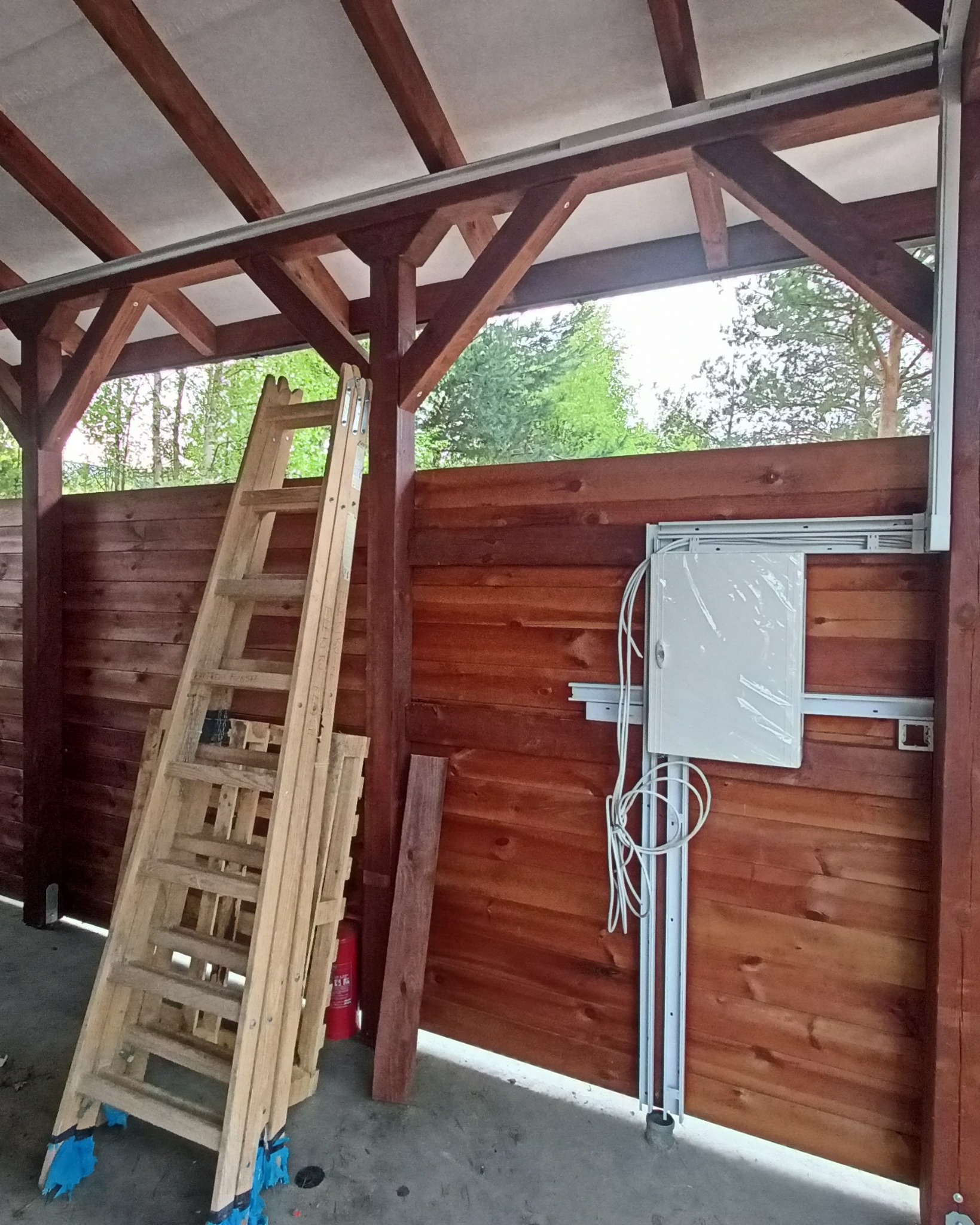 Nowa skrzynka elektryczna z przewodami zamontowana na drewnianej ścianie altany, obok drabina i gaśnica. Widoczne elementy konstrukcji dachu.
