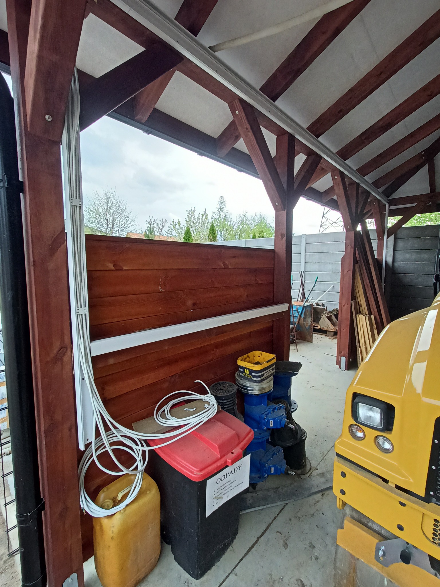 Kable elektryczne prowadzone wzdłuż drewnianej konstrukcji z widoczną maszyną budowlaną w tle, oświetlone naturalnym światłem.
