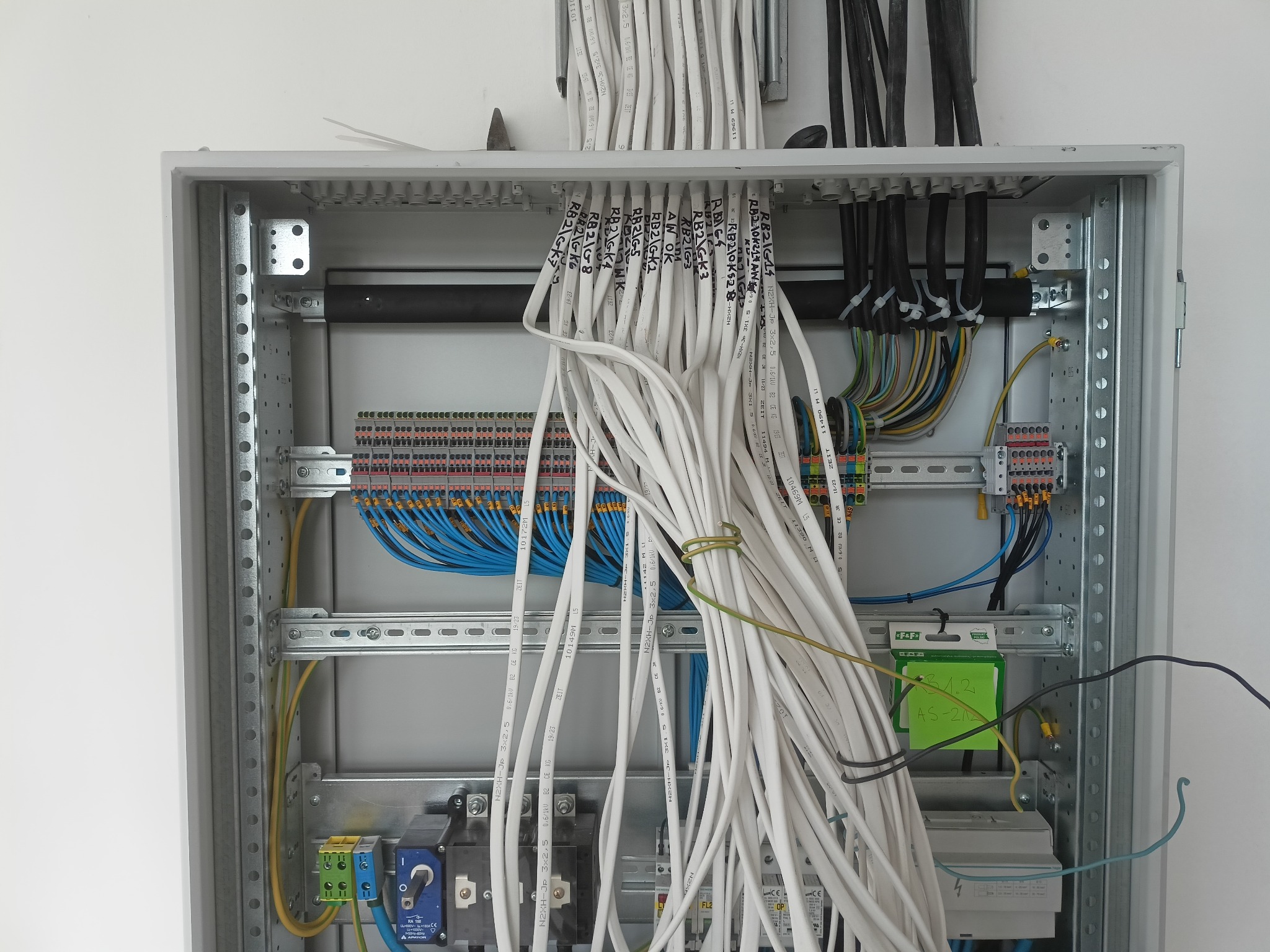 Otwarta skrzynka elektryczna z licznymi przewodami oznaczonymi etykietami, widoczne elementy instalacji i złącza. Instalacja w trakcie montażu, wnętrze skrzynki oświetlone.