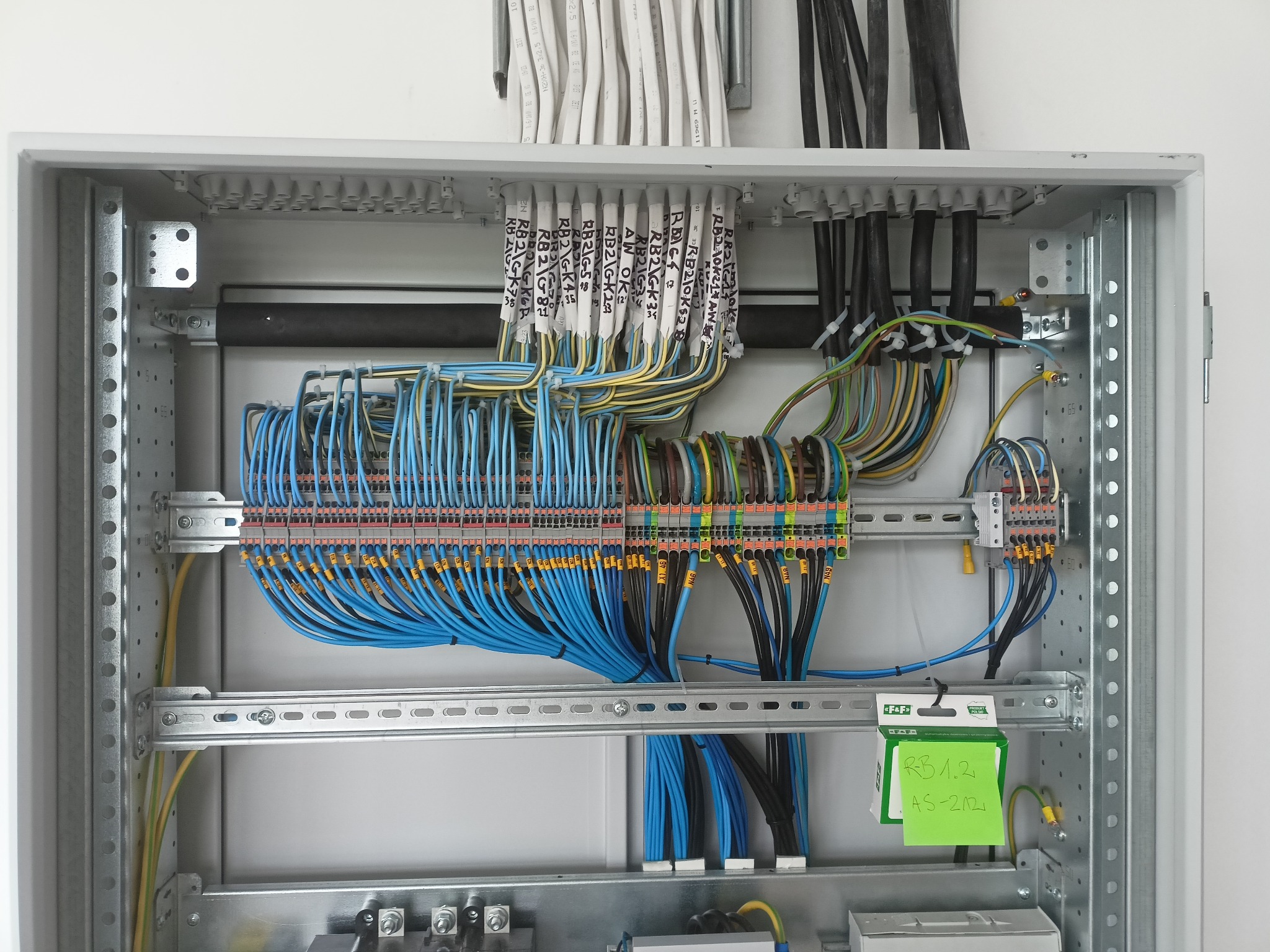 Rozdzielnica elektryczna z precyzyjnie ułożonymi przewodami, oznaczonymi etykietami. Widoczne złącza i elementy montażowe. Na zielonej kartce notatka: RB 1.2, AS-212.