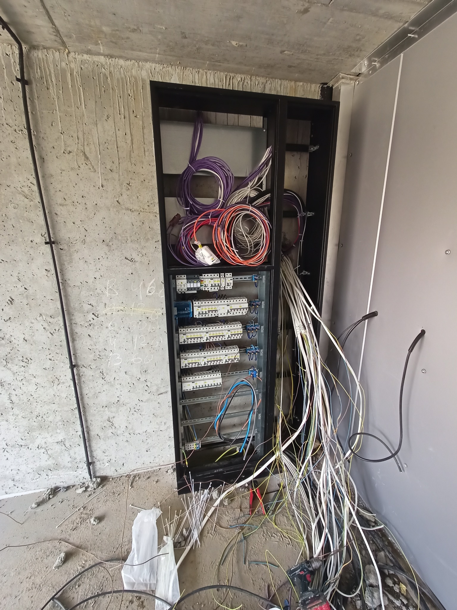 Rozdzielnica elektryczna w trakcie montażu w surowym wnętrzu. Widoczne liczne kable, bezpieczniki i elementy instalacji. Ściany w stanie deweloperskim.