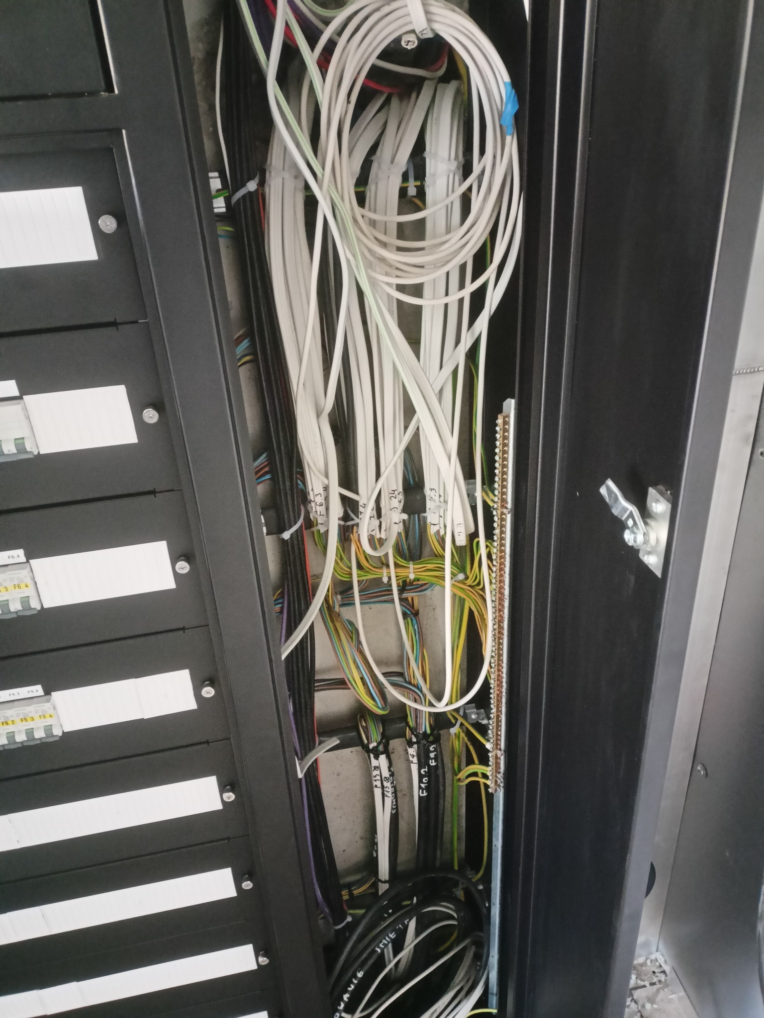 Otwarta szafa elektryczna z widocznymi przewodami, listwami zaciskowymi i bezpiecznikami. Instalacja w trakcie lub po wykonaniu, z podpisami na przewodach.