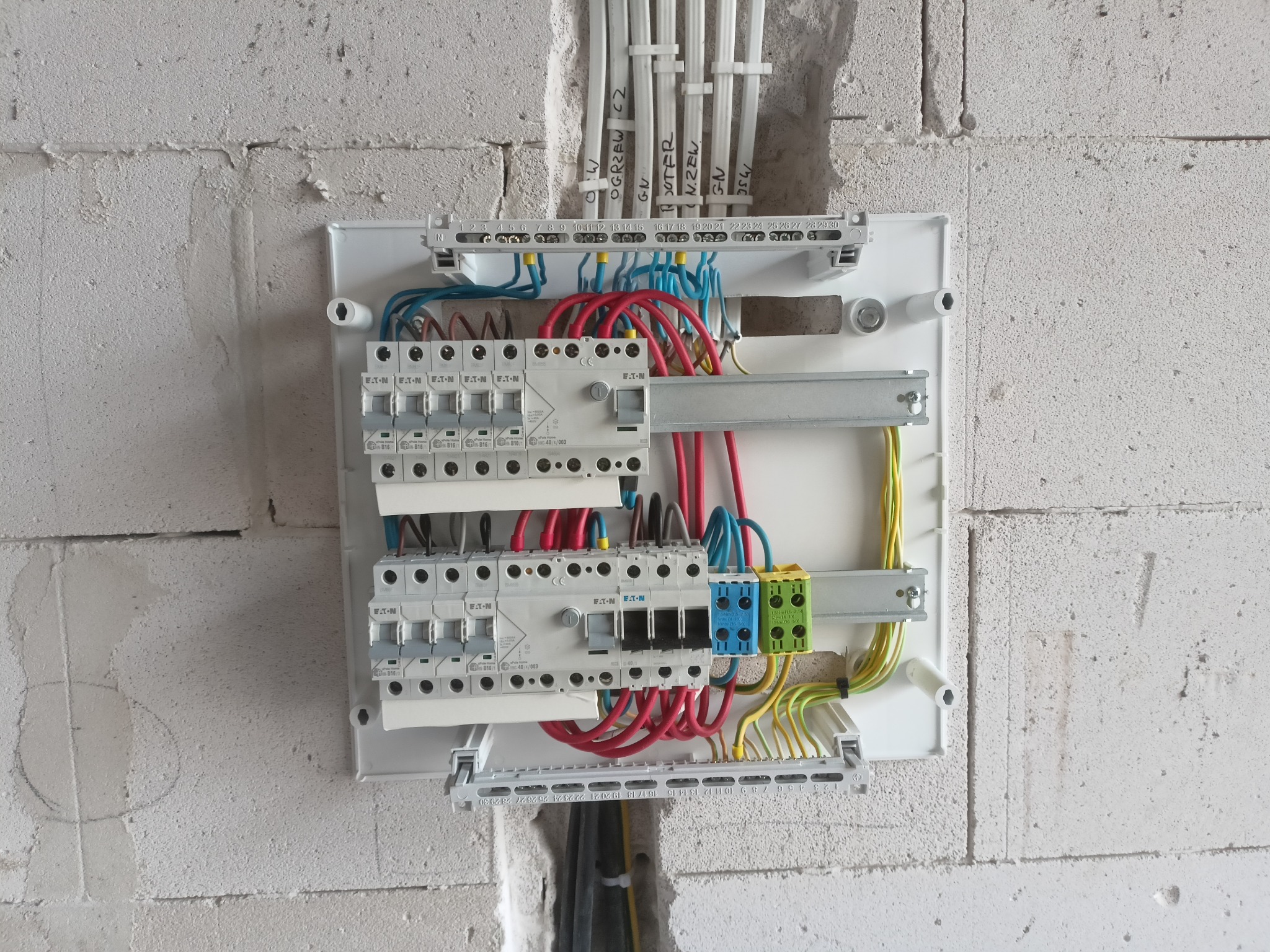 Rozdzielnica elektryczna w trakcie montażu w surowym wnętrzu. Widoczne kable, bezpieczniki EATON i oznaczenia. Ściana z betonu komórkowego.